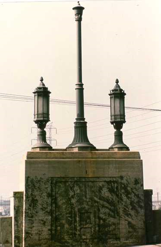 A concrete bridge pylon supporting a tall central pole flanked by two lower lantern fixtures.