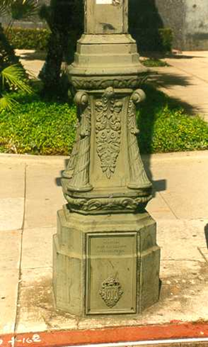 Front-facing close-up of a decorative lamp post base showing a detailed crest and the year 1910 embossed on the metal access door.