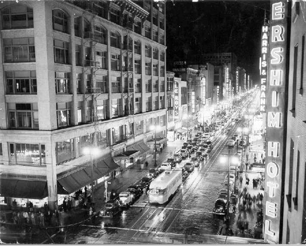 Black and white photo of a busy Los Angeles street at night with neon signs, cars, and bright street lamps.
