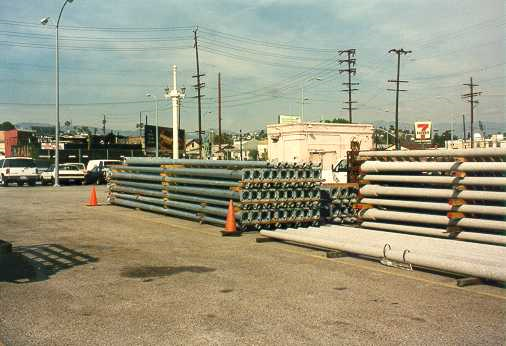 Bundles of straight metal light poles stacked in a paved storage area under a clear sky.