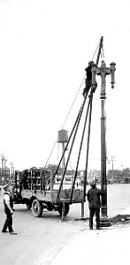 Historic black and white photo of workers using a tripod crane on a truck to install a decorative lamp post.