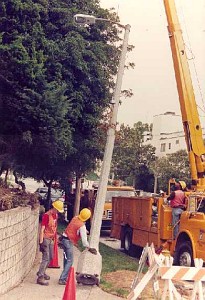 Maintenance crew in hard hats and safety vests using a yellow crane to guide a tall concrete light pole into place.