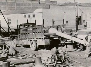 Black and white photo of a flatbed trailer loaded with large concrete light poles in a construction yard.