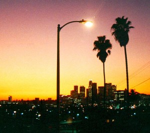 Silhouette of a cobra head streetlight and palm trees against a golden sunset and the Los Angeles skyline.