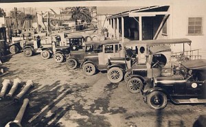 A row of antique open-cab utility trucks parked in a maintenance yard, shown in a sepia-toned vintage photo.