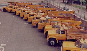 An aerial view of a row of modern yellow flatbed and utility trucks parked in a paved city lot.