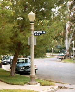 An acorn-style street lamp standing next to a street name sign on a lush, tree-lined residential corner.